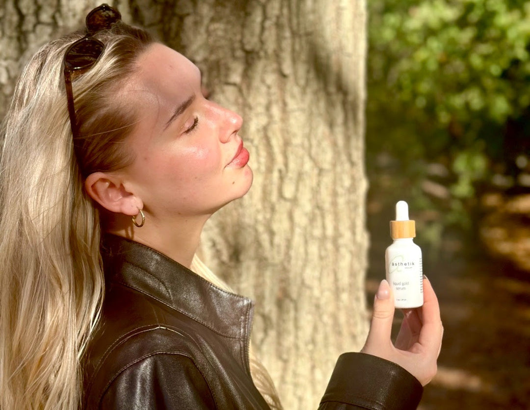 Young blonde woman in a leather jacket standing by a tree with her eyes closed holding a bottle of ästhetik skincare liquid gold face serum