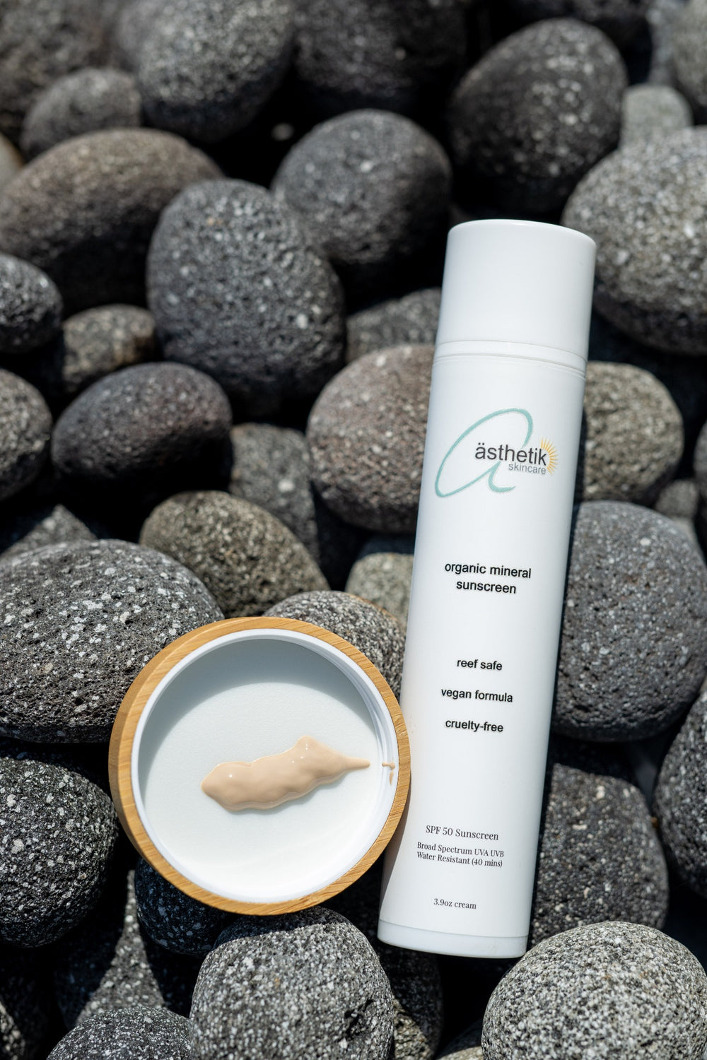 A bottle of ästhetik skincare organic tinted mineral sunscreen SPF 50 next to an open container with cream, all resting on a background of smooth grey stones.