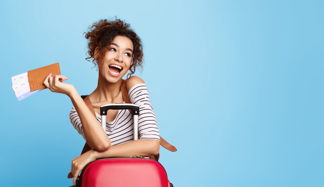 Beautiful young woman with dark curly hair smiling and looking off to the side as she holds tickets and leans against her red suitcase