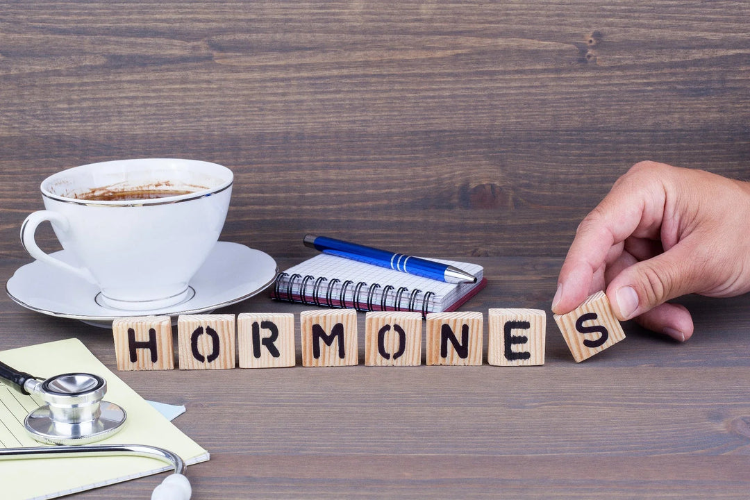 Wooden blocks spelling hormones on a wooden table with a hand placing the last block, next to a stethoscope, notepad, pen, and a cup of coffee