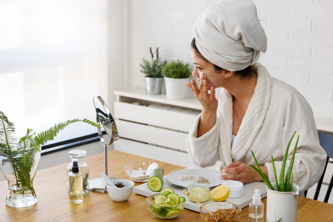 Woman in white robe with towel on head applying face cream, surrounded by natural skincare ingredients and plants
