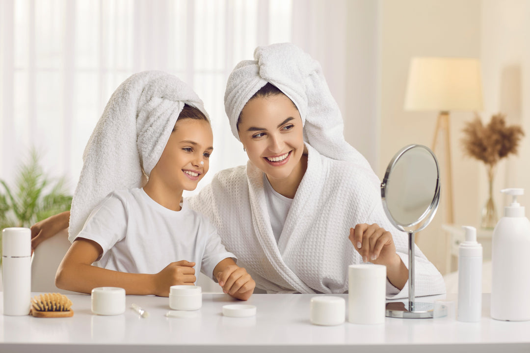 Happy young mother and cute teen daughter enjoy morning beauty treatments at home together. Smiling beautiful family in white bath towels look in cosmetic mirror that stands in front of them on table.