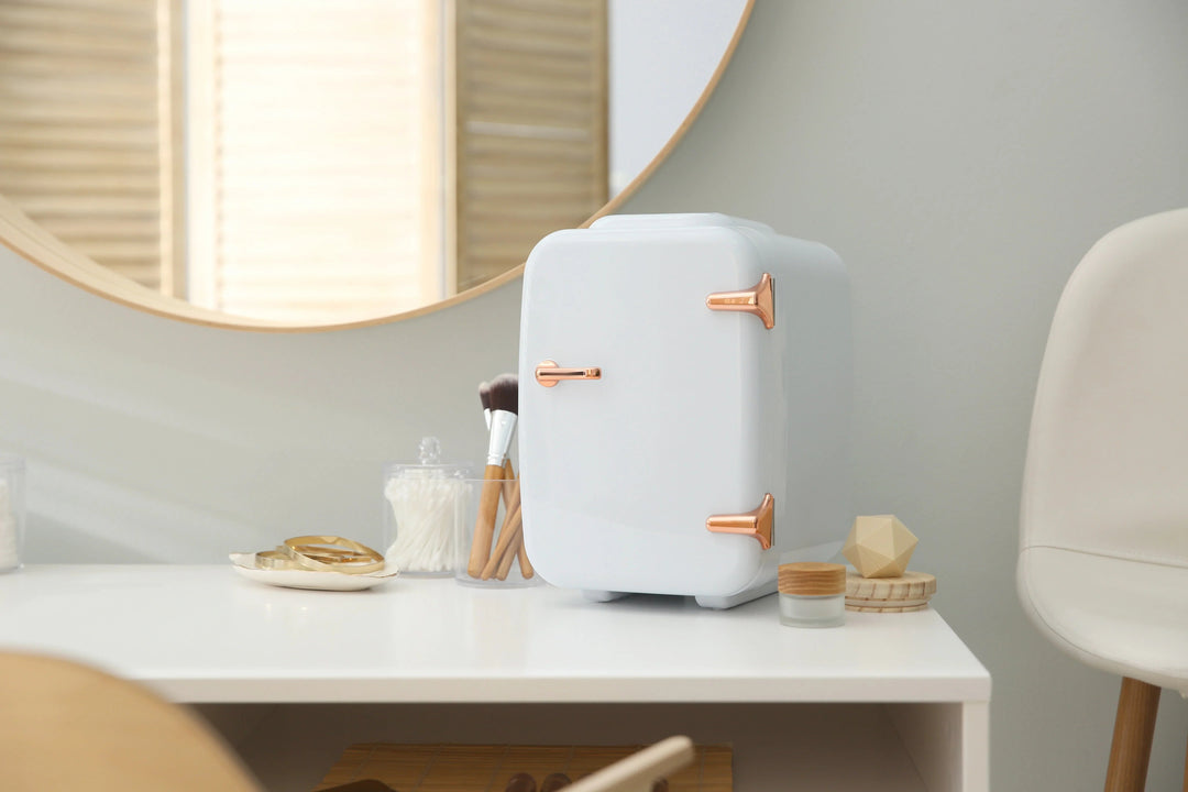 white mini skincare fridge with rose gold handles on white vanity with makeup brushes and cotton swabs