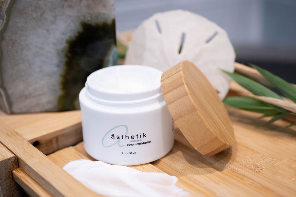 ästhetik skincare ocean moisturizer sitting open on a wooden bath tray with a sand dollar, decorative rock and plants behind it