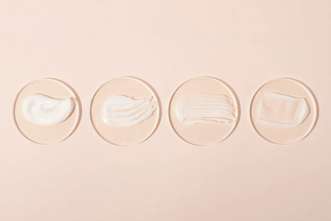 Four different textures of white botanical skincare creams on glass discs against a nude background.