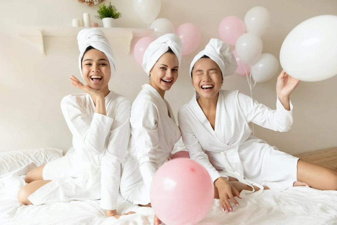 Three happy women in white robes and hair towels celebrating with pink and white balloons.