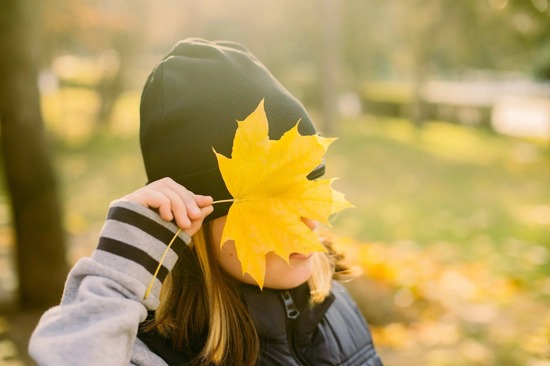 Child wearing a black beanie holding a bright yellow maple leaf over their face in autumn.