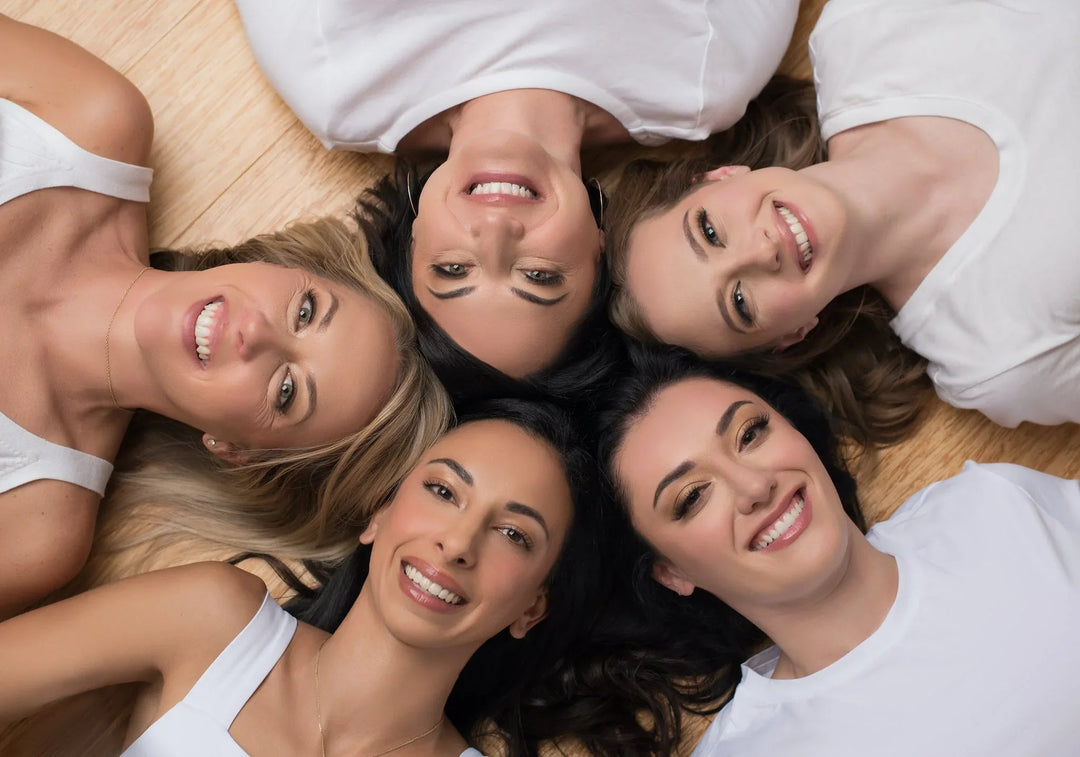 Five smiling diverse women lying on wooden floor in white tops, showcasing radiant, healthy skin for skincare brand