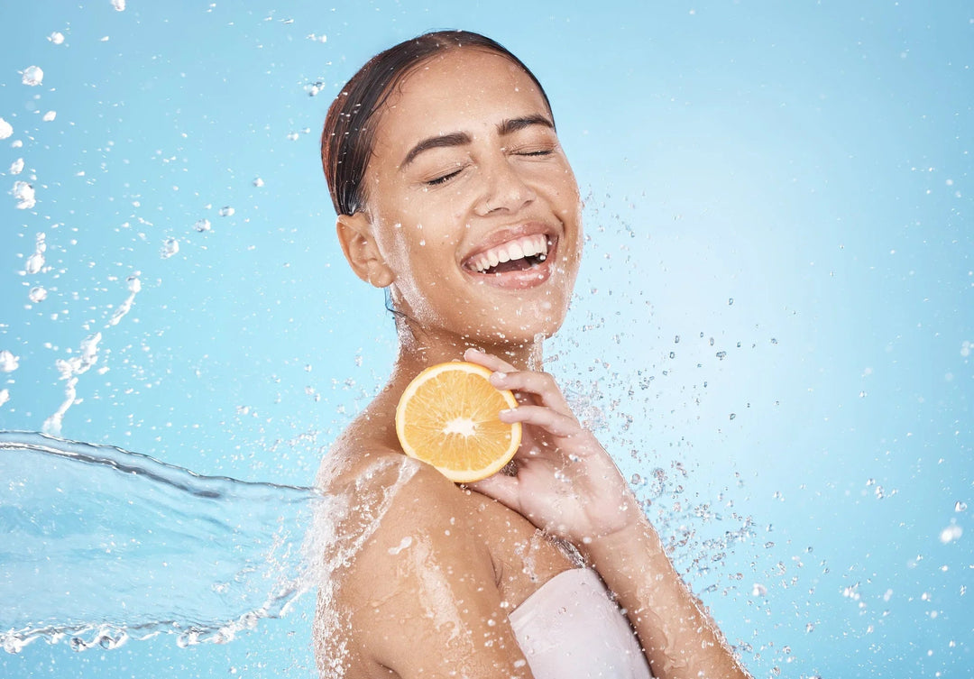 Smiling woman with glowing skin holding an orange slice amidst splashing water