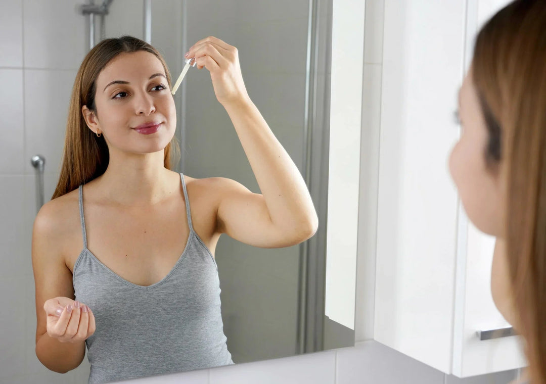 Woman applying skincare serum with dropper in bathroom mirror, promoting healthy skin routine