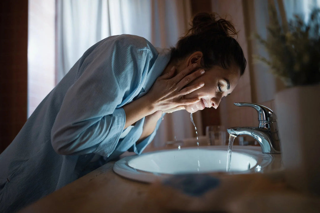 Woman washing her face with water at a bathroom sink for a clean beauty skincare routine.