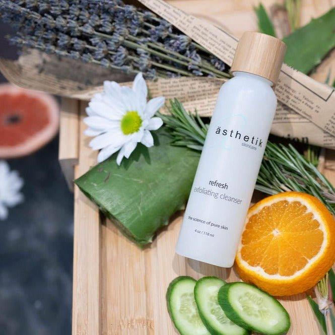 asthetik skincare refresh exfoliating cleanser on a wooden bath board with citus fruits, aloe, rosemary, cucumber, lavender and flowers surrounding it