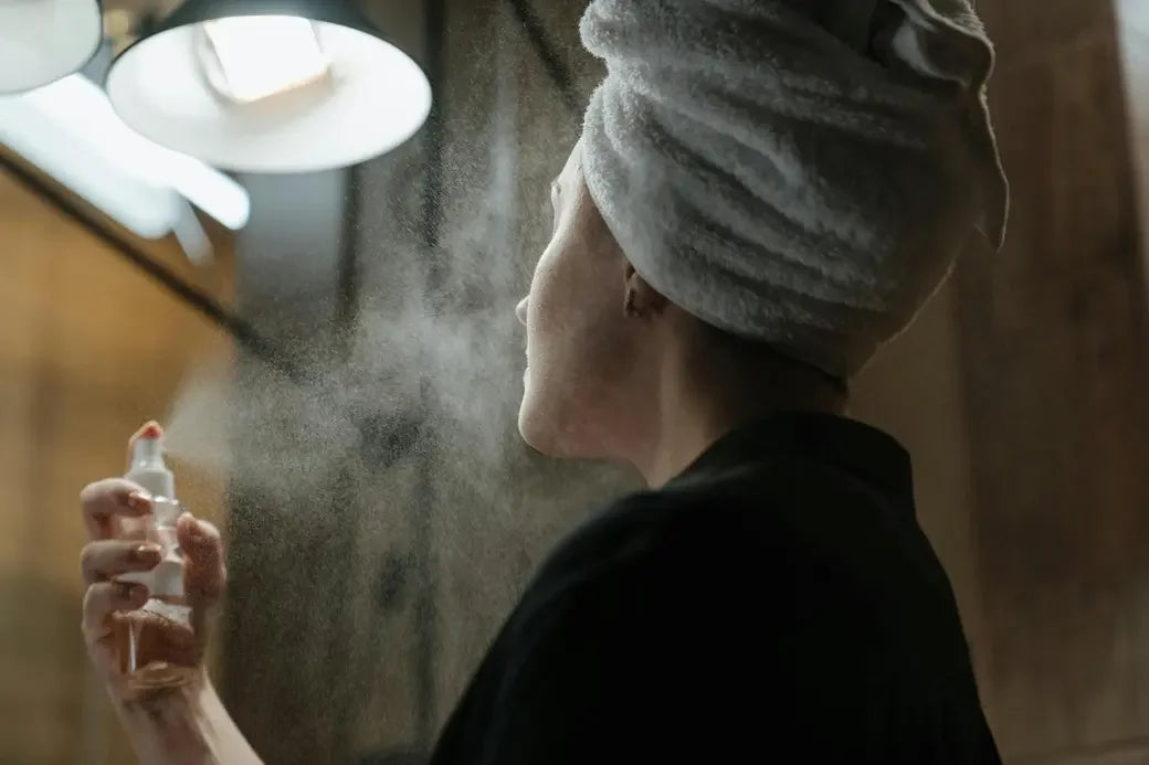 A woman wearing a white head wrap using a skincare facial mist spray.