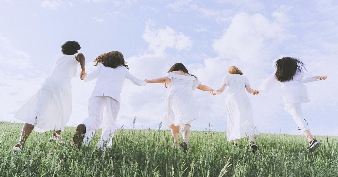 Five diverse women in white clothing holding hands and walking through a green grassy field.