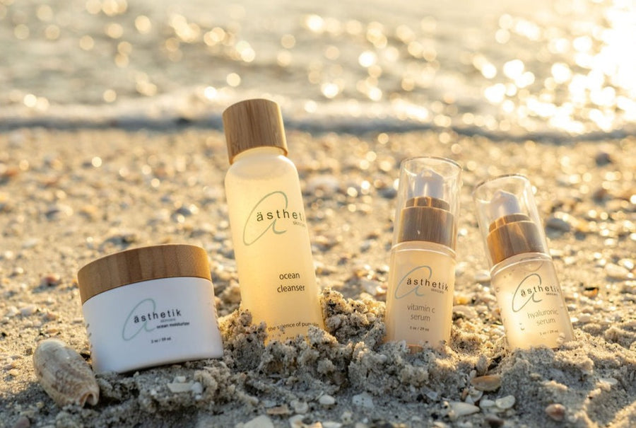Sunset on a beach with a lineup of skincare products from the ästhetik skincare ocean collection arranged in the sand, featuring bottles and jars labeled "astelle.