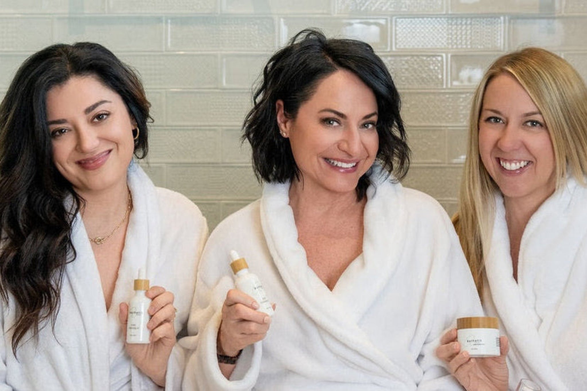 Three women in robes holding asthetik skincare products in a bathroom setting