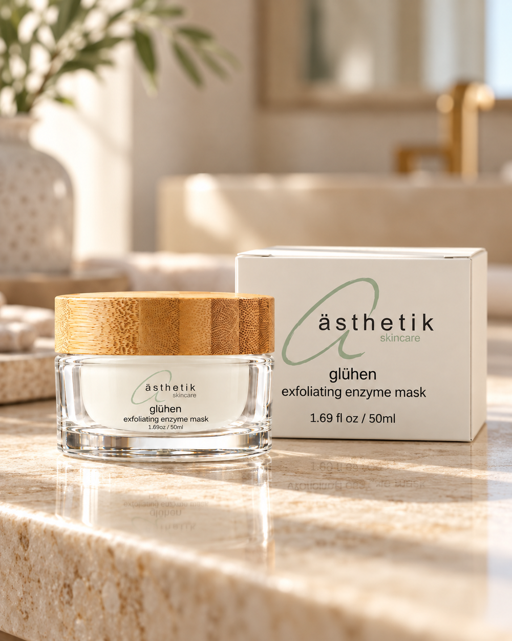 A jar of ästhetik glühen exfoliating enzyme mask with a bamboo-look lid sits on a marble countertop next to its white box, with a softly blurred bathroom background.