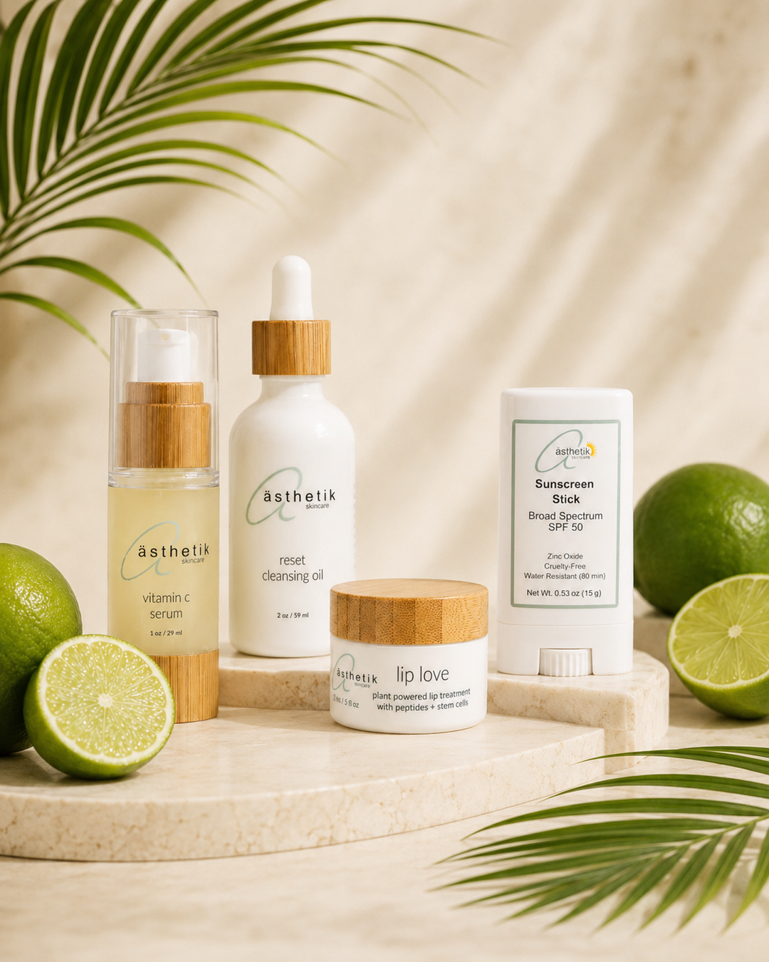 A set of four ästhetik skincare products arranged on a marble tray with lime slices and palm leaves. The products include Vitamin C Serum, Reset cleansing Oil, Lip Love treatment, and Sunscreen Stick.