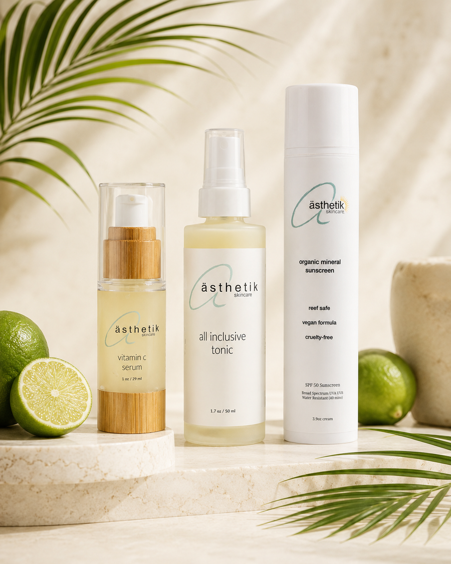 Three ästhetik skincare products arranged on a marble slab with limes and palm leaves: a Vitamin C Serum in a clear bottle with a bamboo-look pump, an All Inclusive Tonic in a frosted bottle with a white spray top, and a mineral Sunscreen in a tall white bottle.