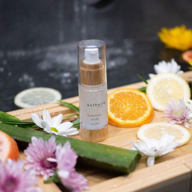 bottle of ästhetik skincare hyaluronic serum with fresh citrus slices, flowers, and leaves on a wooden tray