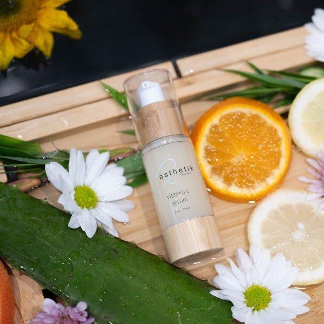 A glass bottle of ästhetik skincare's vitamin C serum resting on a wooden tray, surrounded by fresh citrus slices, green leaves, and vibrant flowers.